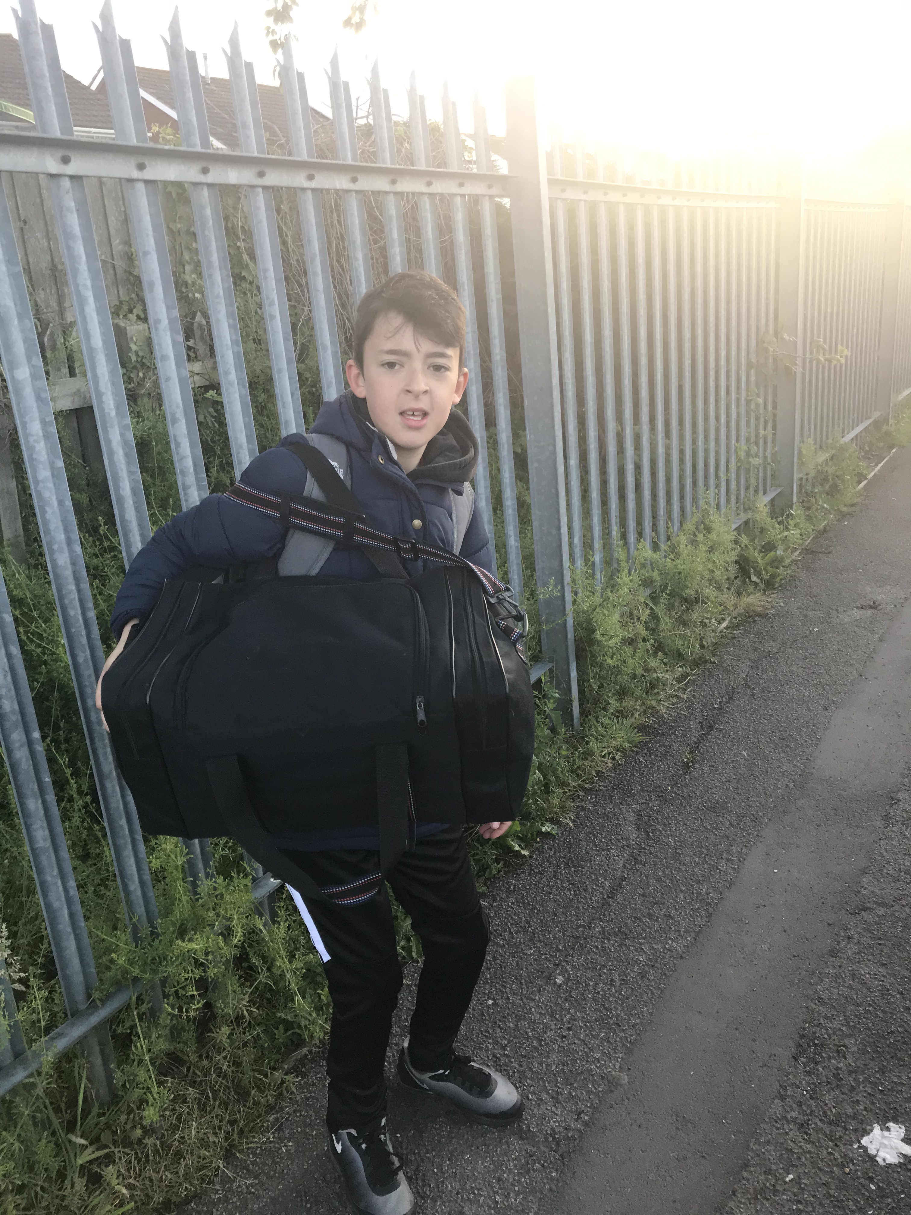 Young boy carrying large bag walking down a street with metal fences