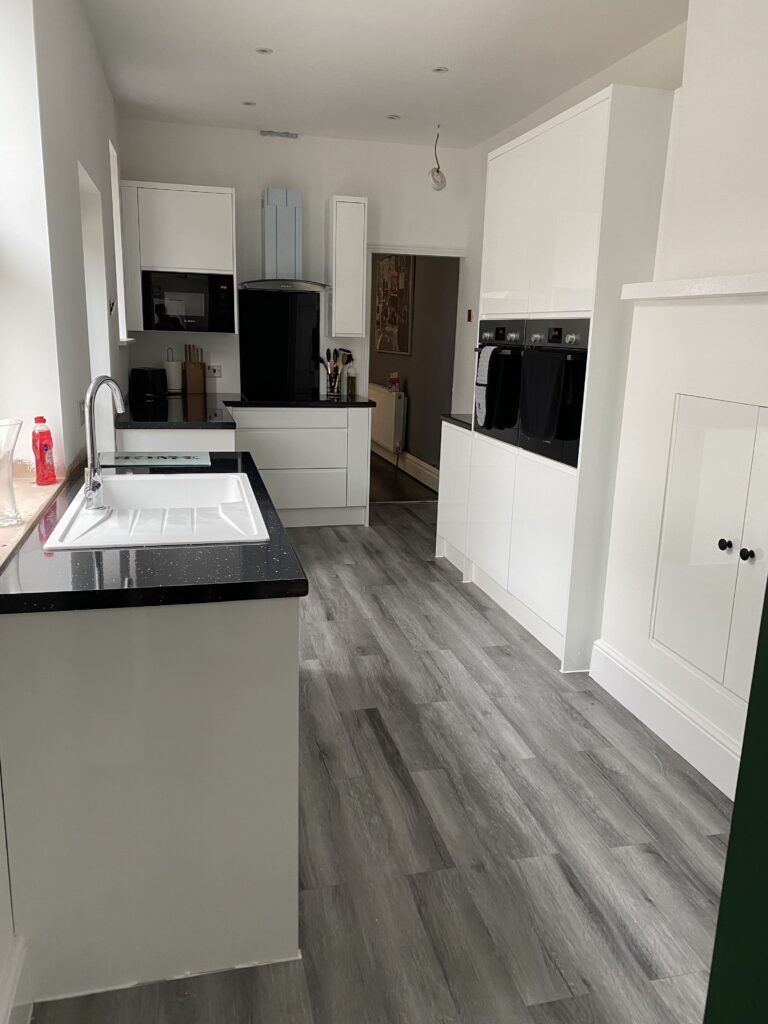 Brand new white kitchen with black gloss worksurfaces, grey wood effect flooring and white and green room